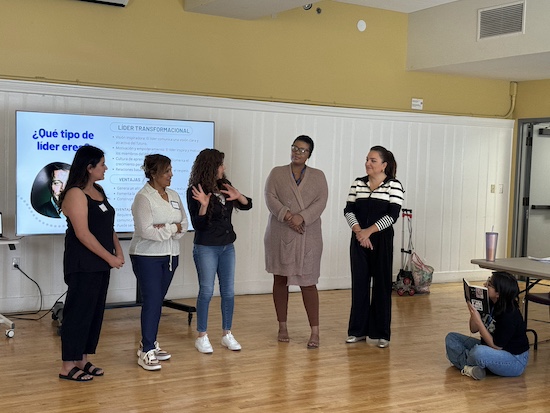 Five women stand in front of a presentation screen during a leadership development workshop. One participant is speaking, illustrating The Women's Building focus on empowering leaders in the community."